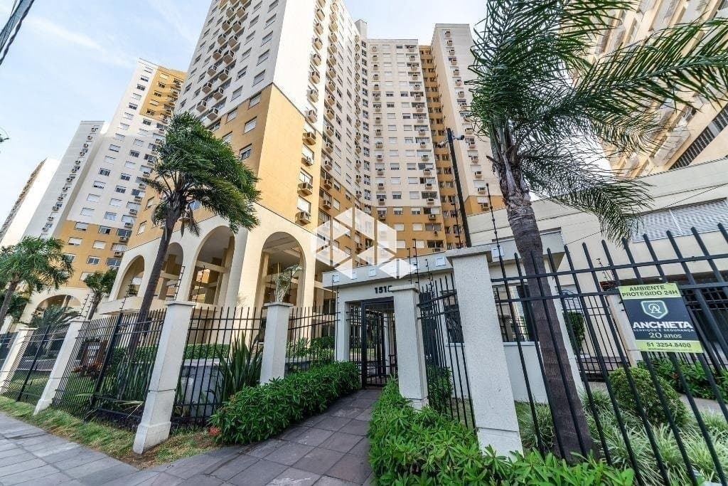 Fachada do edifício residencial alto com portaria, portão de ferro e jardim frontal.